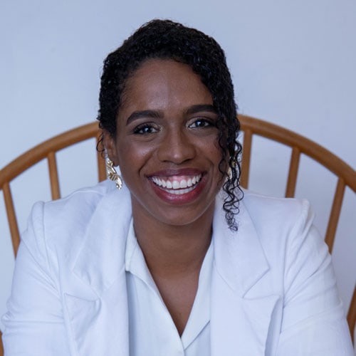 Sorriso de mulher negra com cabelo cacheado e blazer branco em ambiente profissional.